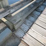 Damaged timber railing at the bridge’s south curb, showing impact deformation, cracked wood, and loose fasteners near the curb edge.