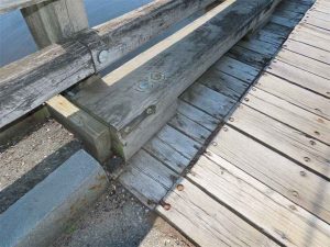 Damaged timber railing at the bridge’s south curb, showing impact deformation, cracked wood, and loose fasteners near the curb edge.