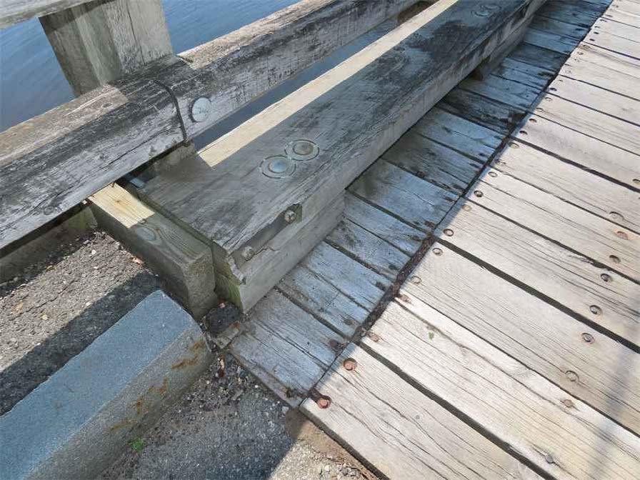 Damaged timber railing at the bridge’s south curb, showing impact deformation, cracked wood, and loose fasteners near the curb edge.