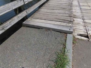 Transition between the asphalt sidewalk and timber bridge showing vertical displacement and vegetation growth at the edge, indicating settlement.