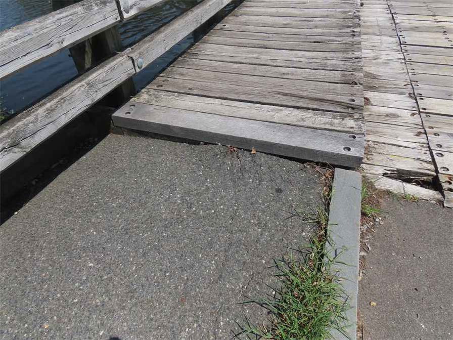 Transition between the asphalt sidewalk and timber bridge showing vertical displacement and vegetation growth at the edge, indicating settlement.