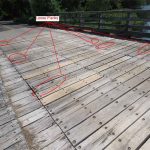 Midspan view of timber bridge deck looking east, showing weathered planks with signs of wear, discoloration, and minor splitting along the surface. Six portions are circled and labelled as loose planks.